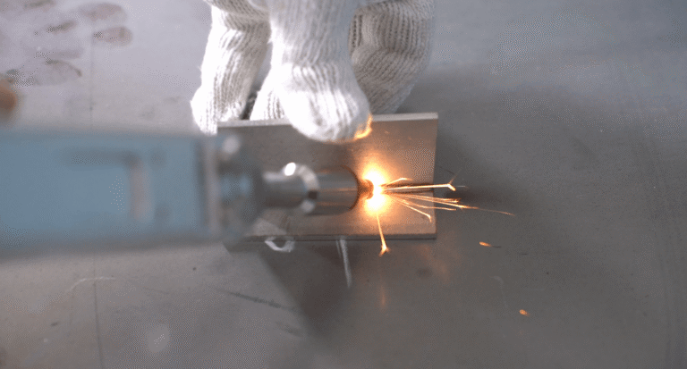 Una mano con un guanto bianco regge una piastra metallica mentre una saldatrice laser portatile emette scintille durante il processo di saldatura.